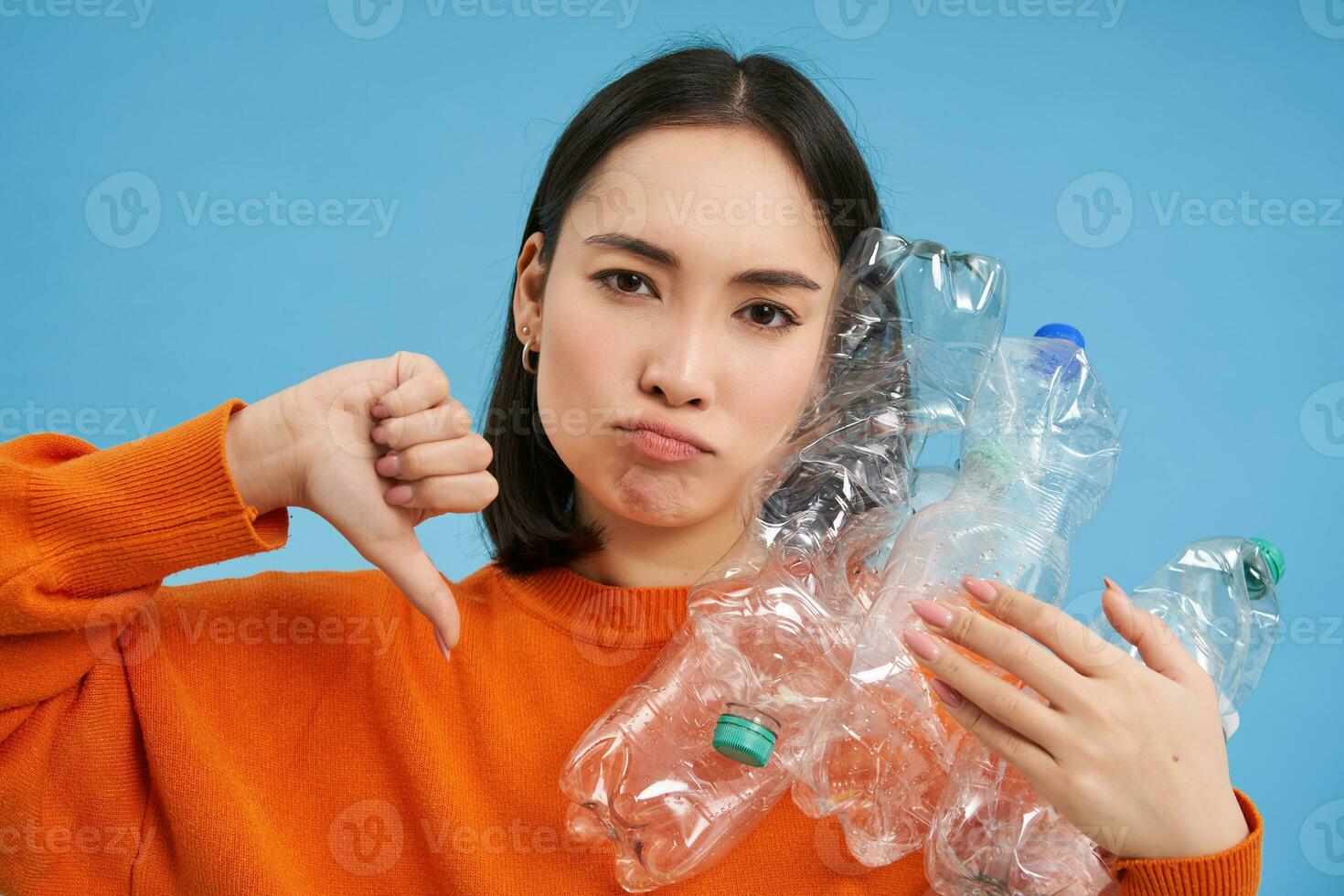 Portrait of woman with upset face, shows thumb down and plastic bottles, disappointed with lack of recycling, blue background photo