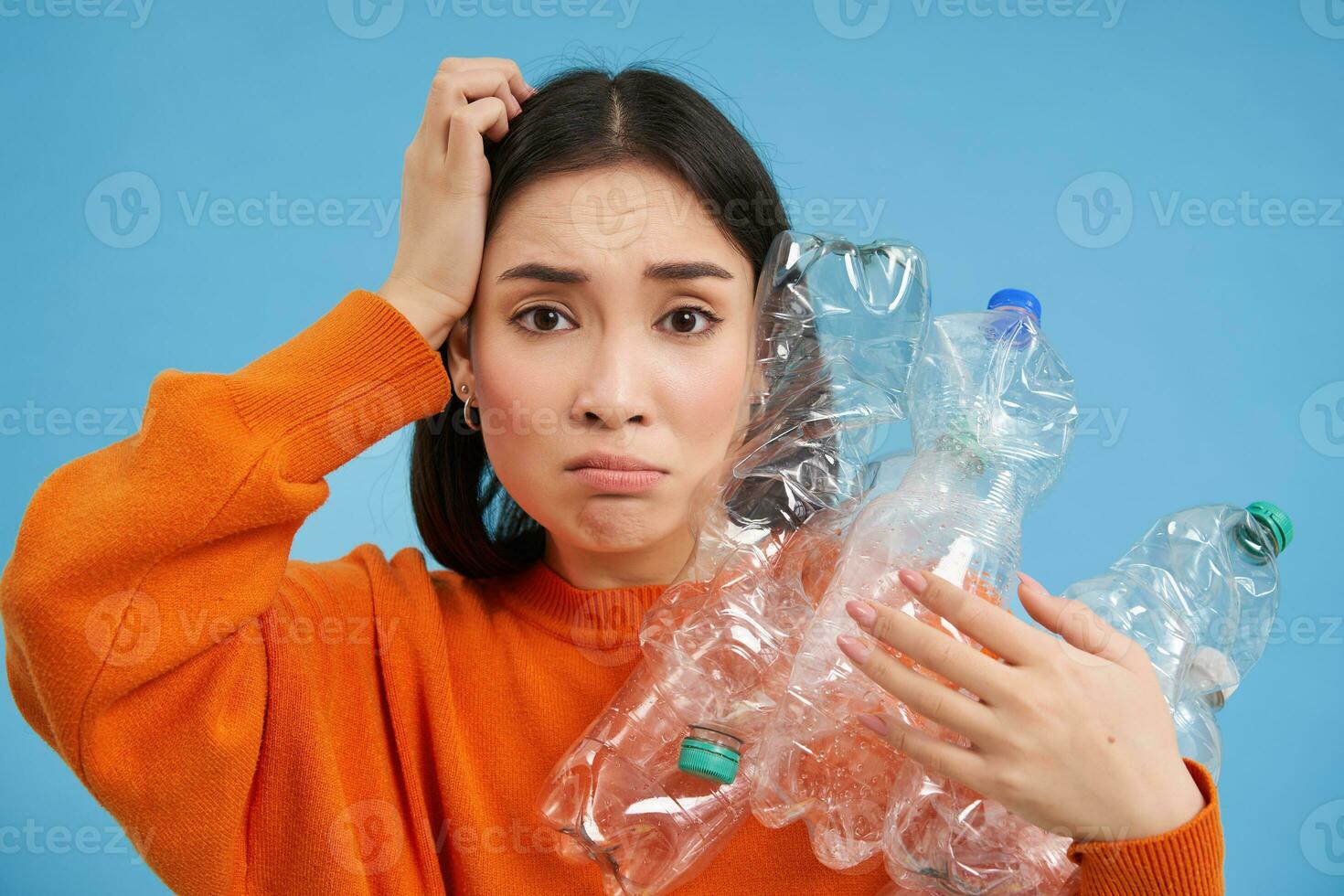 Sad asian woman holding plastic bottles, looking troubled and upset, sorting waste for recycling, standing over blue background photo