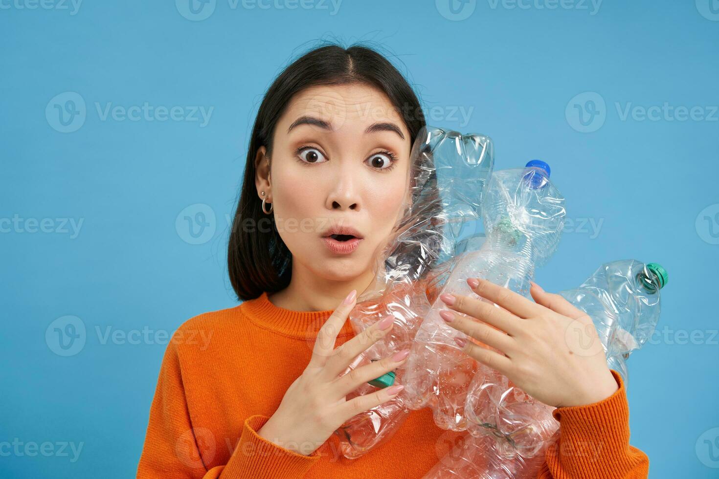 Portrait of cute korean woman, holding two plastic bottles with excited face, recycling her waste, sorting garbage, blue background photo