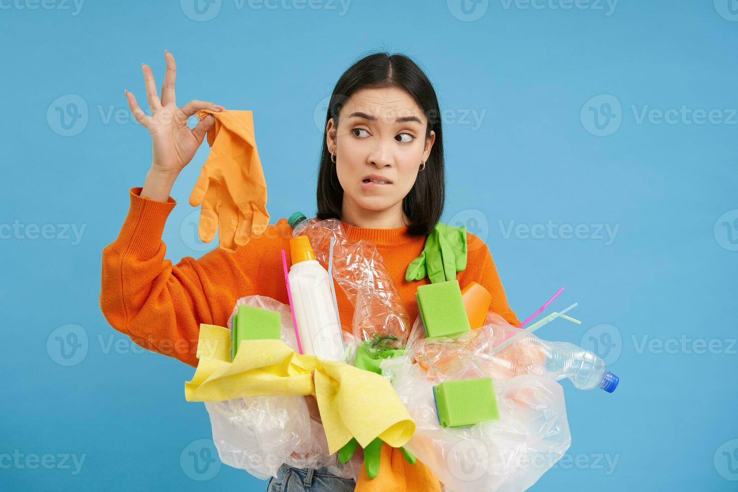 Young woman holds plastic garbage, looks at glove with shocked face, stinky waste, sorting litter for recycling, blue background photo