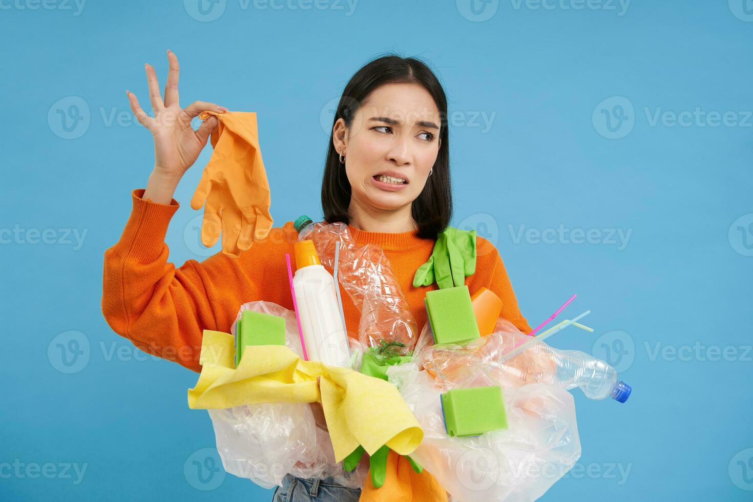 Korean woman looks at stinky latex glove, holds plastic garbage, cleans house and recycle, blue background photo