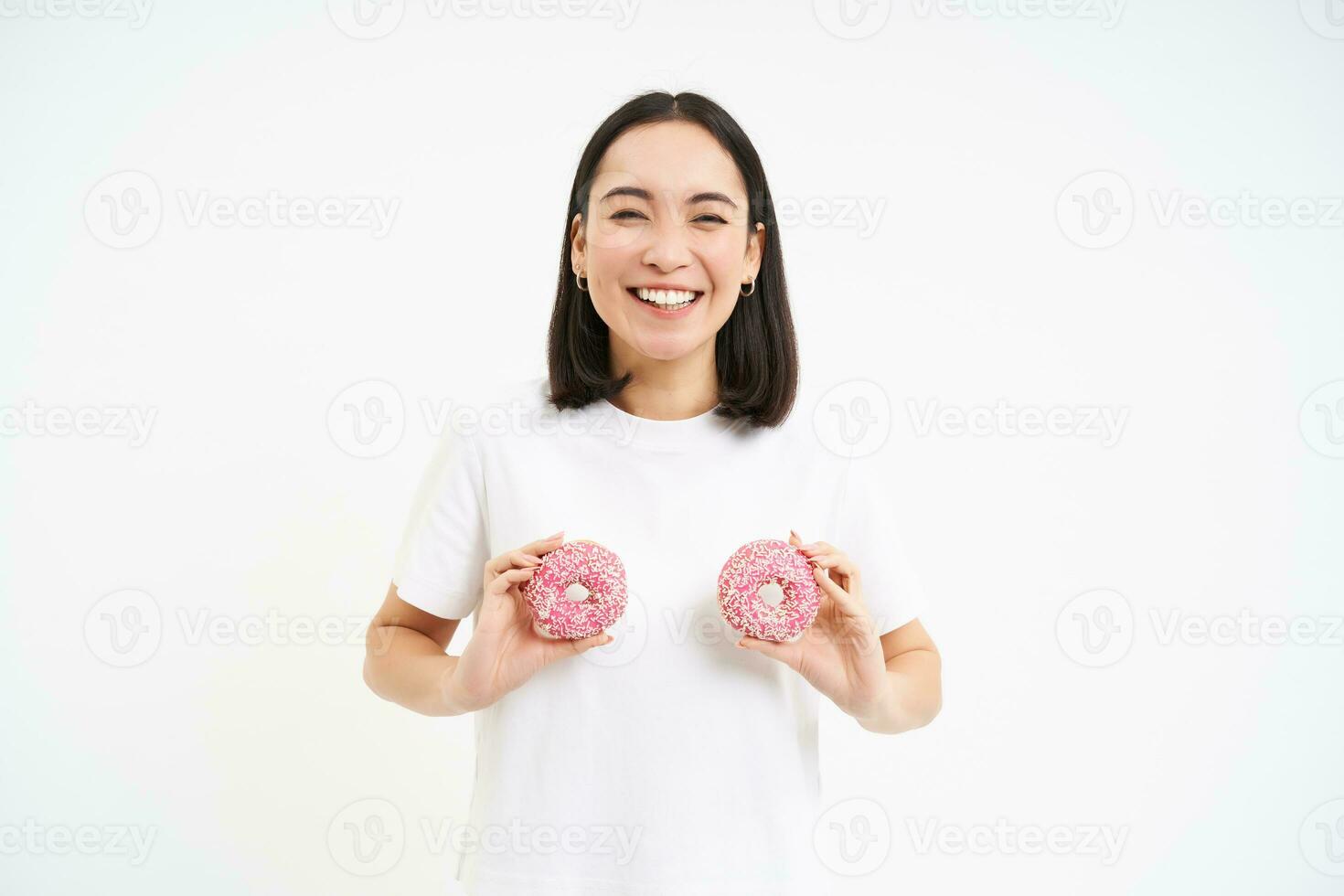 Funny korean woman posing with two delicious glazed doughnuts on chest, laughing and smiling, eats junk food, tasty dessert photo