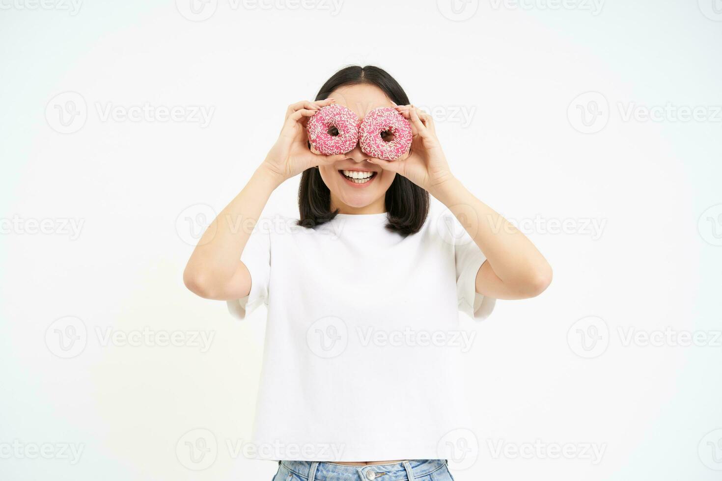 Portrait of happy korean woman, makes glasses from two tasty glazed doughnuts, smiling and having fun, white background photo