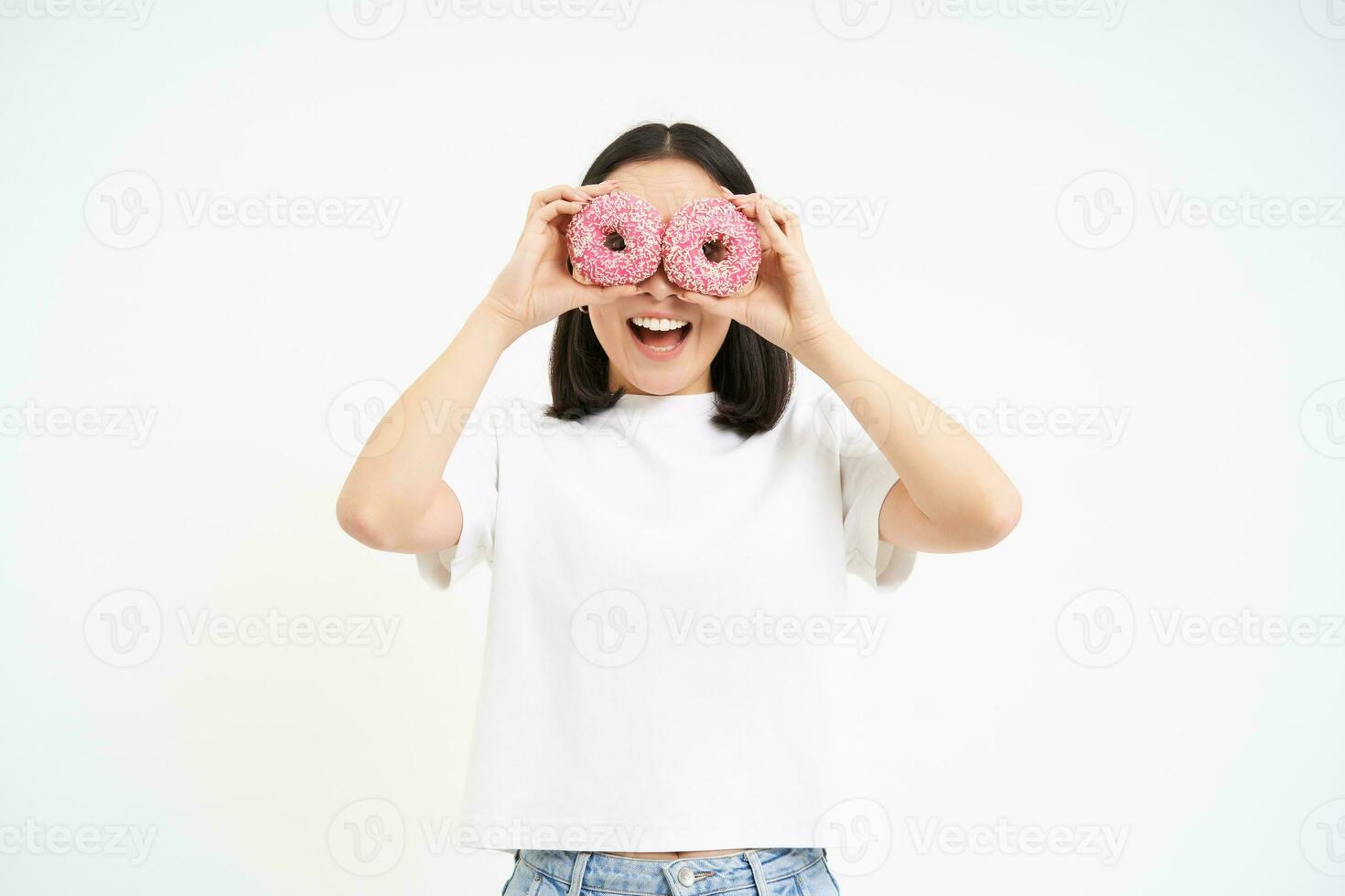 Funny smiling korean woman, holding two glazed pink doughnuts, eating dessert with happy face, white background photo