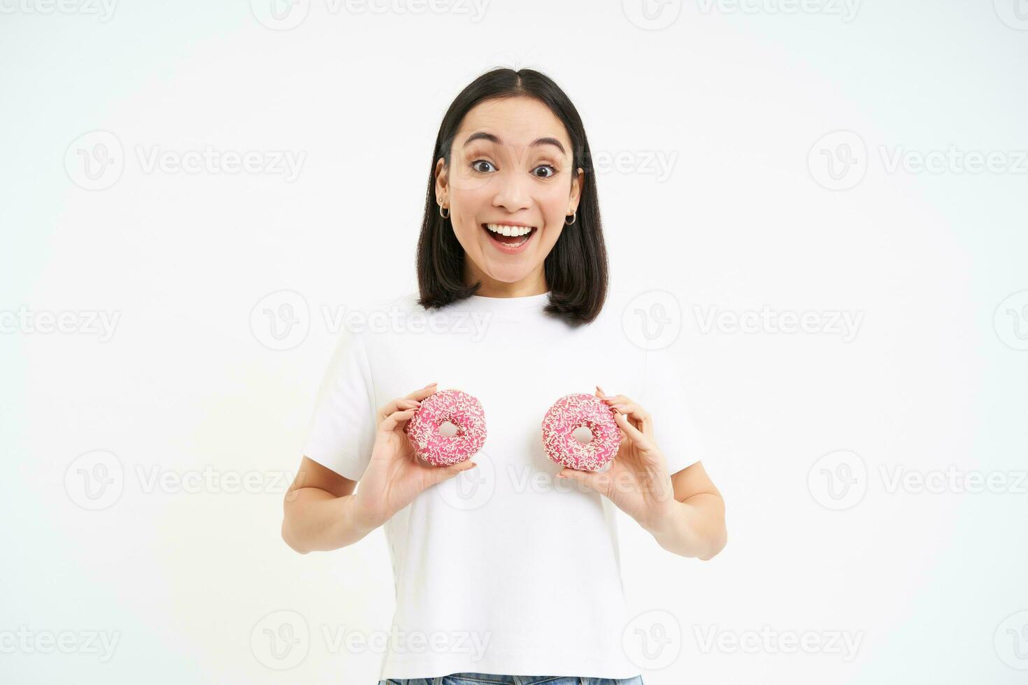 Funny korean woman posing with two delicious glazed doughnuts on chest, laughing and smiling, eats junk food, tasty dessert photo