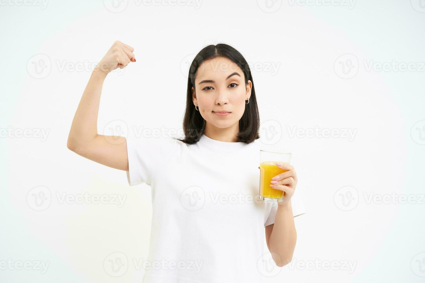 Healthy lifestyle. Smiling young asian woman, shows strong muscles, biceps on hand and drinks orange juice, white background photo