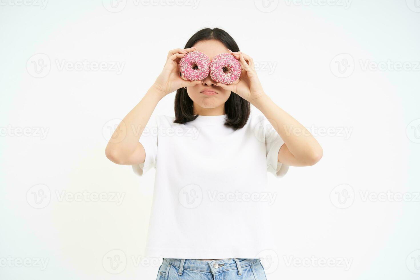 Image of funny young woman, makes glasses with two pink glazed doughnuts, looking through donnut holes and smiling, isolated on white background photo