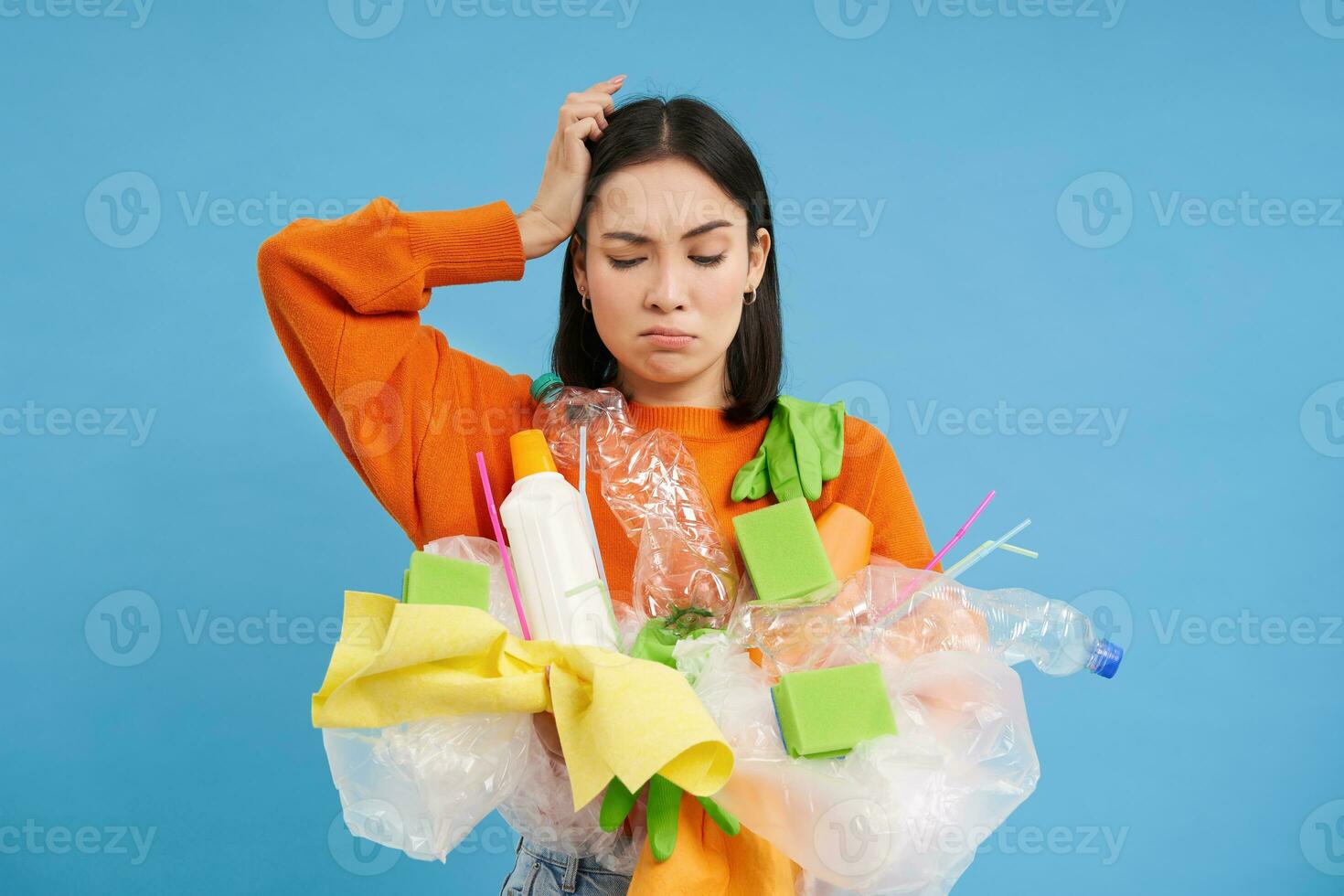 Confused Korean woman tries to go green, recycle plastic household items, holding bottles and gloves, does recycling, blue background photo