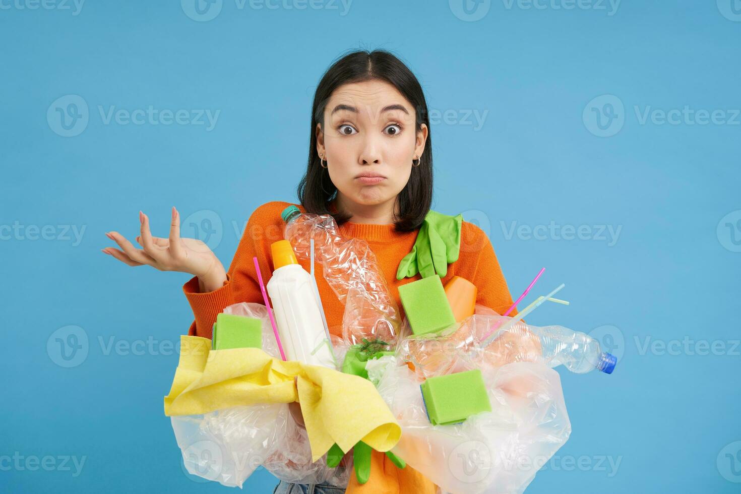 Korean girl with plastic garbage, shrugs shoulders, doesnt know how to recycle, sorting her waste for recycling station, blue background photo