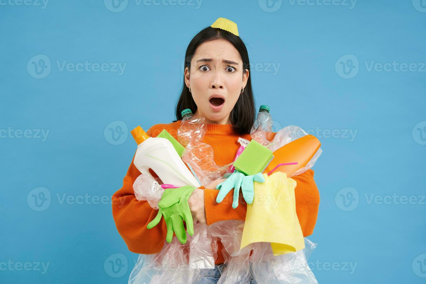 Sad asian woman holding garbage, empty plastic bottles and waste, tired of recycling, sorting rubbish, isolated on blue background photo