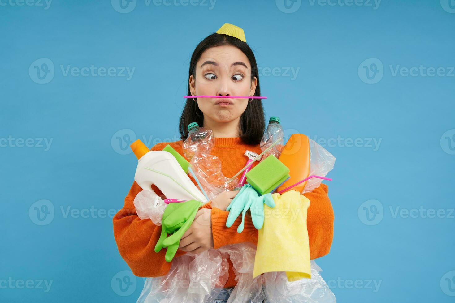 Funny asian woman holding plastic garbage and makes faces, collecting waste for recycling center, blue background photo
