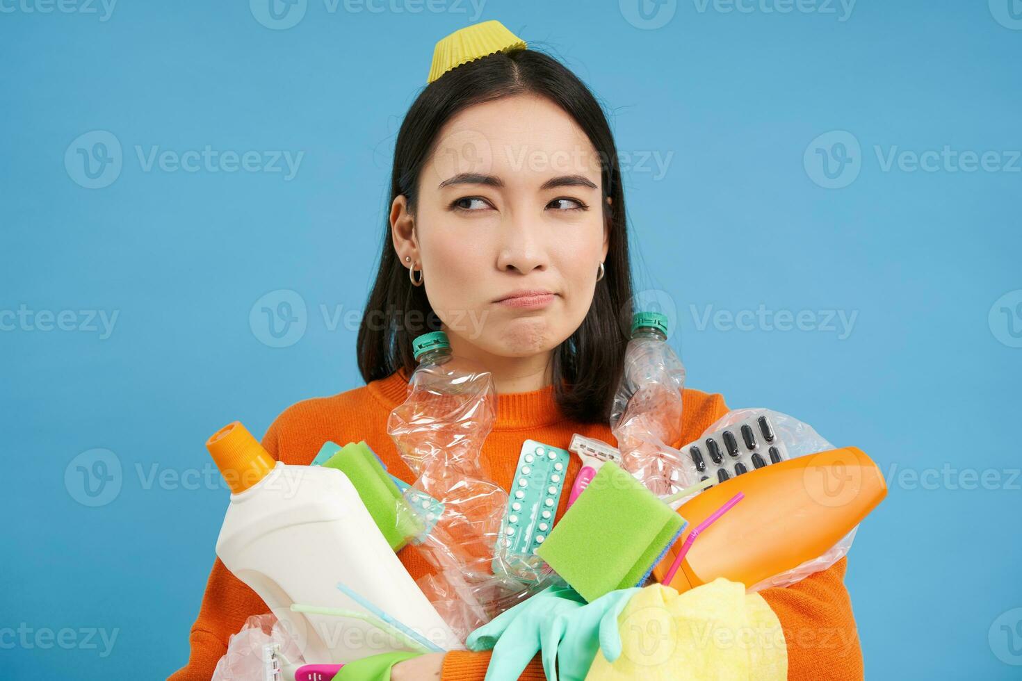 Puzzled young woman with pile of garbage, holds plastic bottles and trash for recycling, looks perplexed, blue background photo