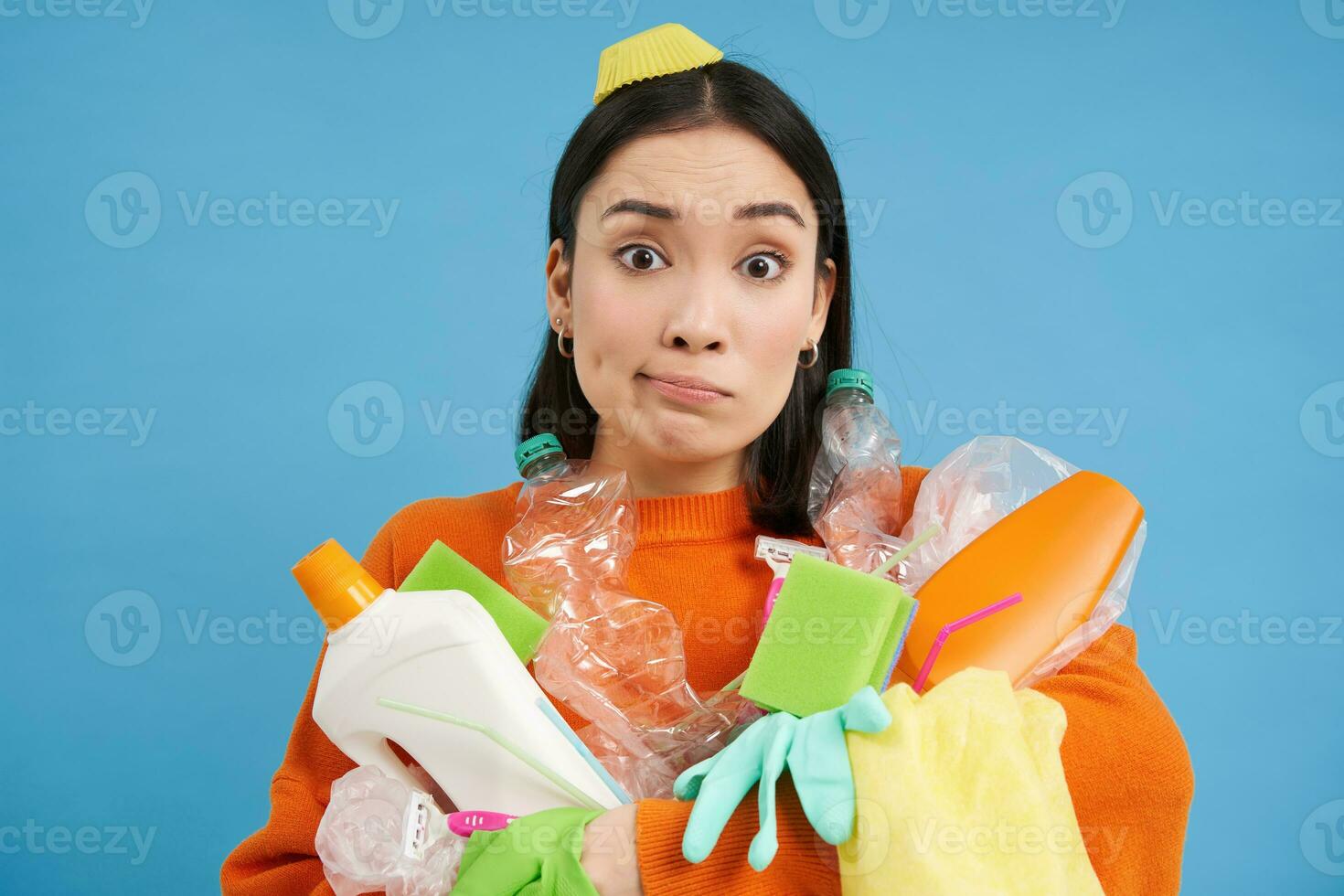 Puzzled young woman with pile of garbage, holds plastic bottles and trash for recycling, looks perplexed, blue background photo