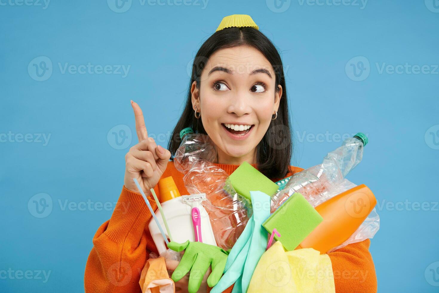 Enthusiastic asian woman, holding plastic empty bottles and garbage for sorting and recycling, pointing finger up at recycle center, blue background photo