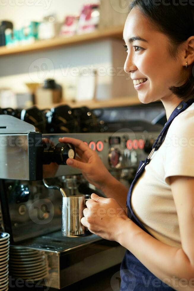 Happy asian woman, barista using coffee machine to make order, steaming milk for cappuccino and latte, laughing and smiling while working in cafe photo