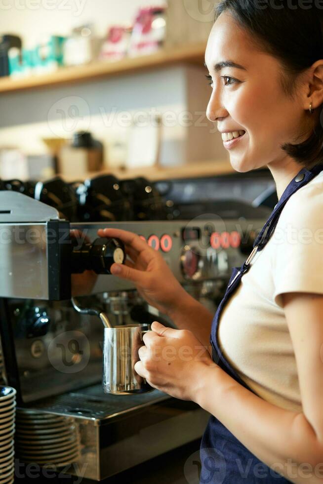 contento asiático mujer, barista utilizando café máquina a hacer orden, humeante Leche para capuchino y latté, riendo y sonriente mientras trabajando en café foto