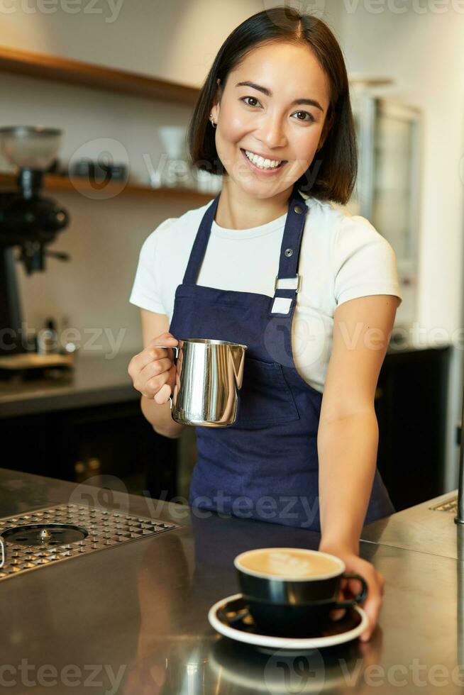 Vertical shot of smiling asian girl in cafe, giving out cappuccino, made order, prepared coffee for you, standing behind counter in blue apron photo