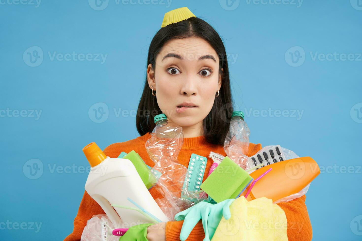Portrait of asian woman with intrigued face, holds rubbish for recycling, empty plastic bottles and waste, stares confused, blue background photo
