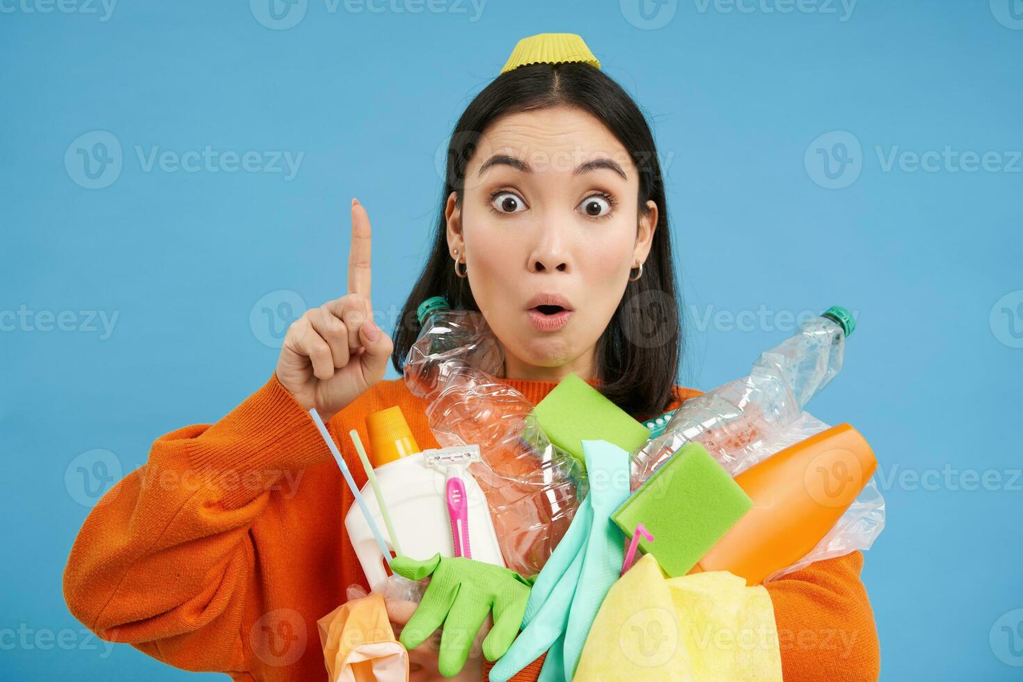 Close up portrait of asian woman pointing up, showing banner, holding recycling items, empty plastic bottles, sorting garbage, isolated on blue background photo