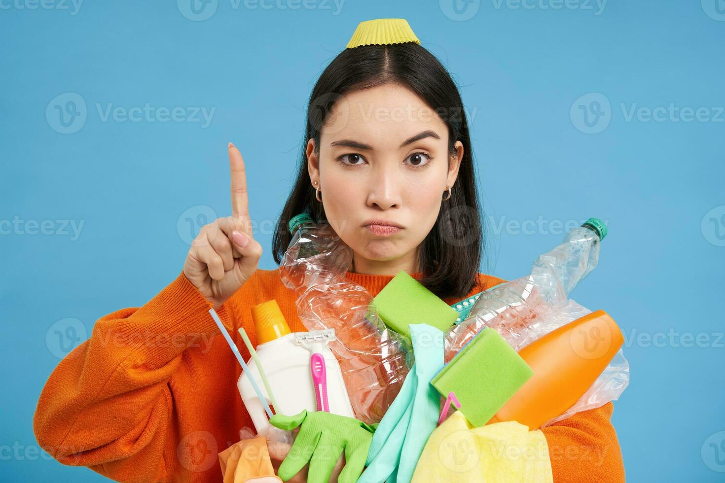 Close up portrait of asian woman pointing up, showing banner, holding recycling items, empty plastic bottles, sorting garbage, isolated on blue background photo