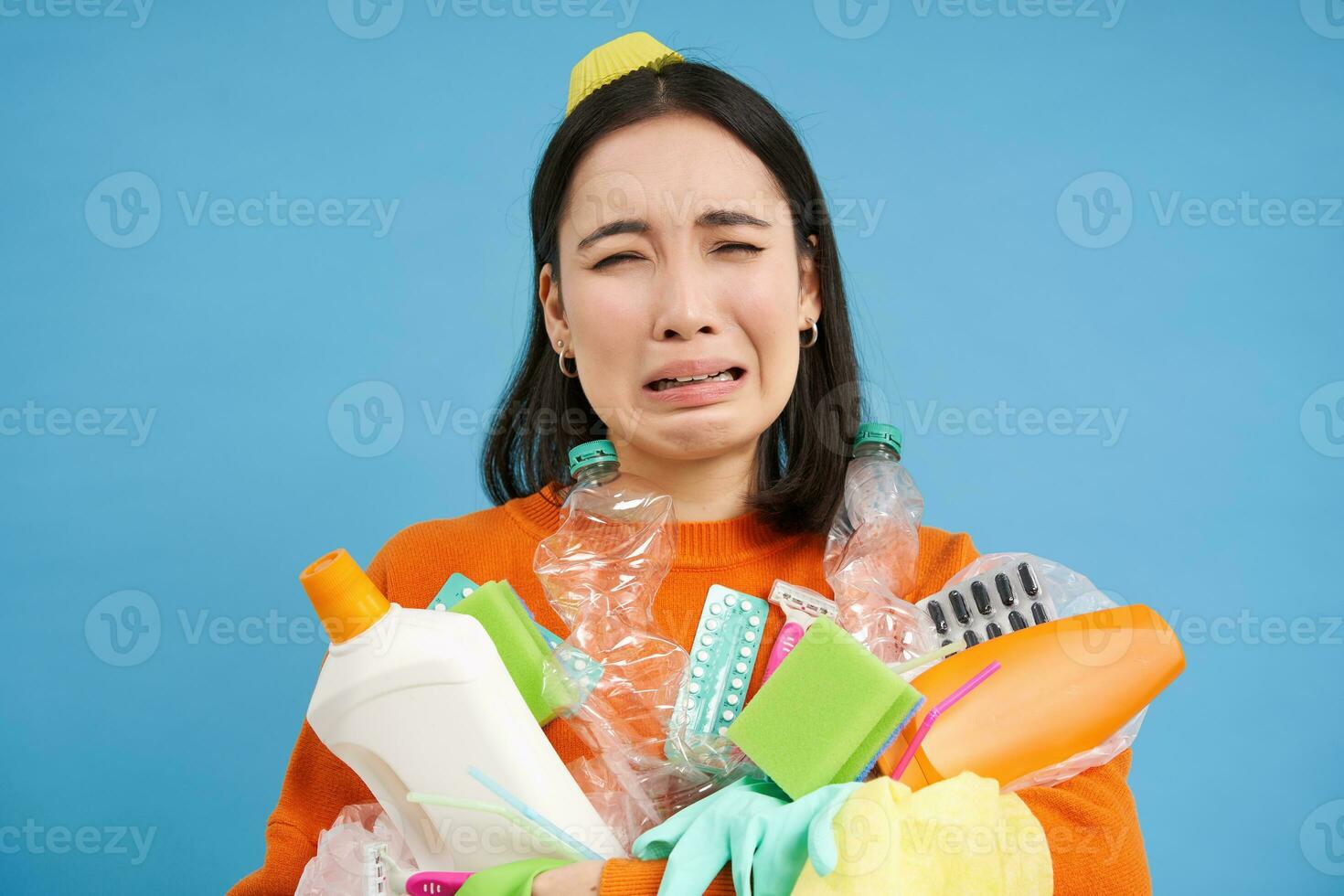 Portrait of crying sad girl with plastic trash, collects recycling waste, looks miserable, blue background photo