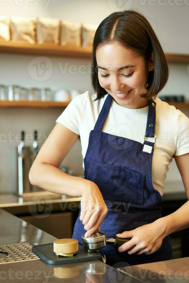 Vertical shot of barista, asian girl in cafe using tamper for coffee machine, wearing blue apron, preparing order photo