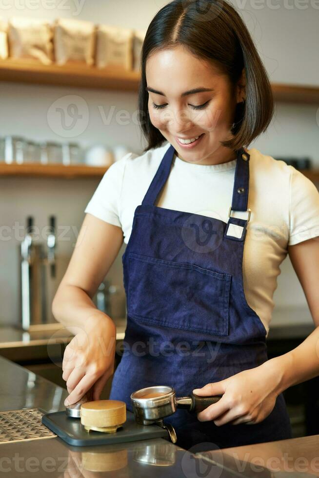 Vertical shot of smiling asian girl putting coffee in tamper to brew capuccino, prepare order in coffee machine, standing behind counter in blue apron photo