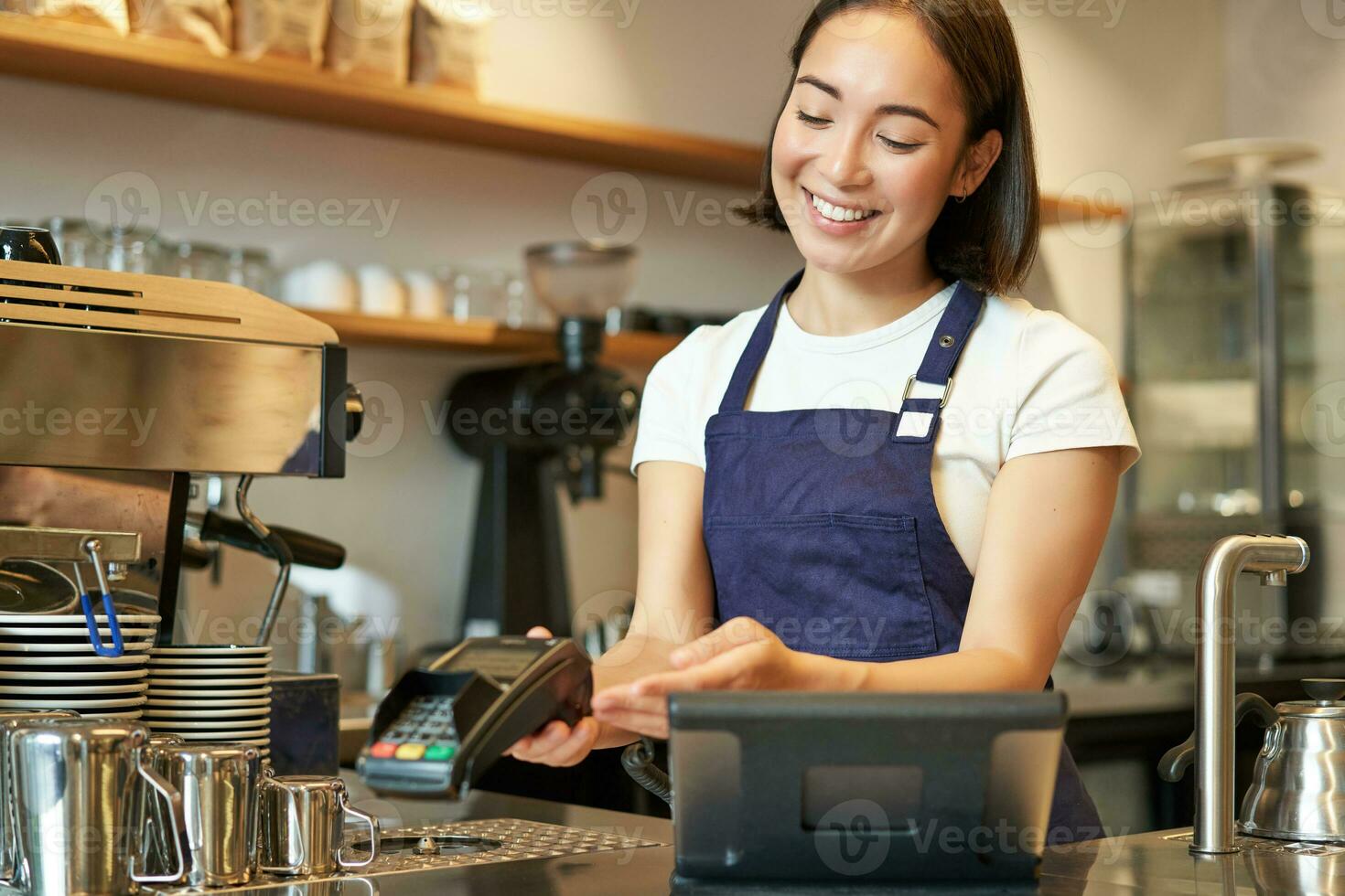 Smiling asian girl barista, giving client POS terminal, help pay with credit card, standing at counter in coffee shop, working in cafe photo