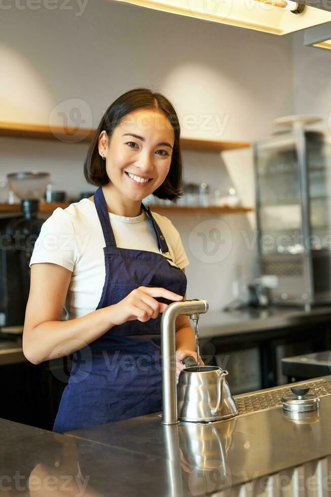 Vertical shot of barista, girl in coffee shop, pouring water in kettle, preparing order in cafe, smiling at camera photo