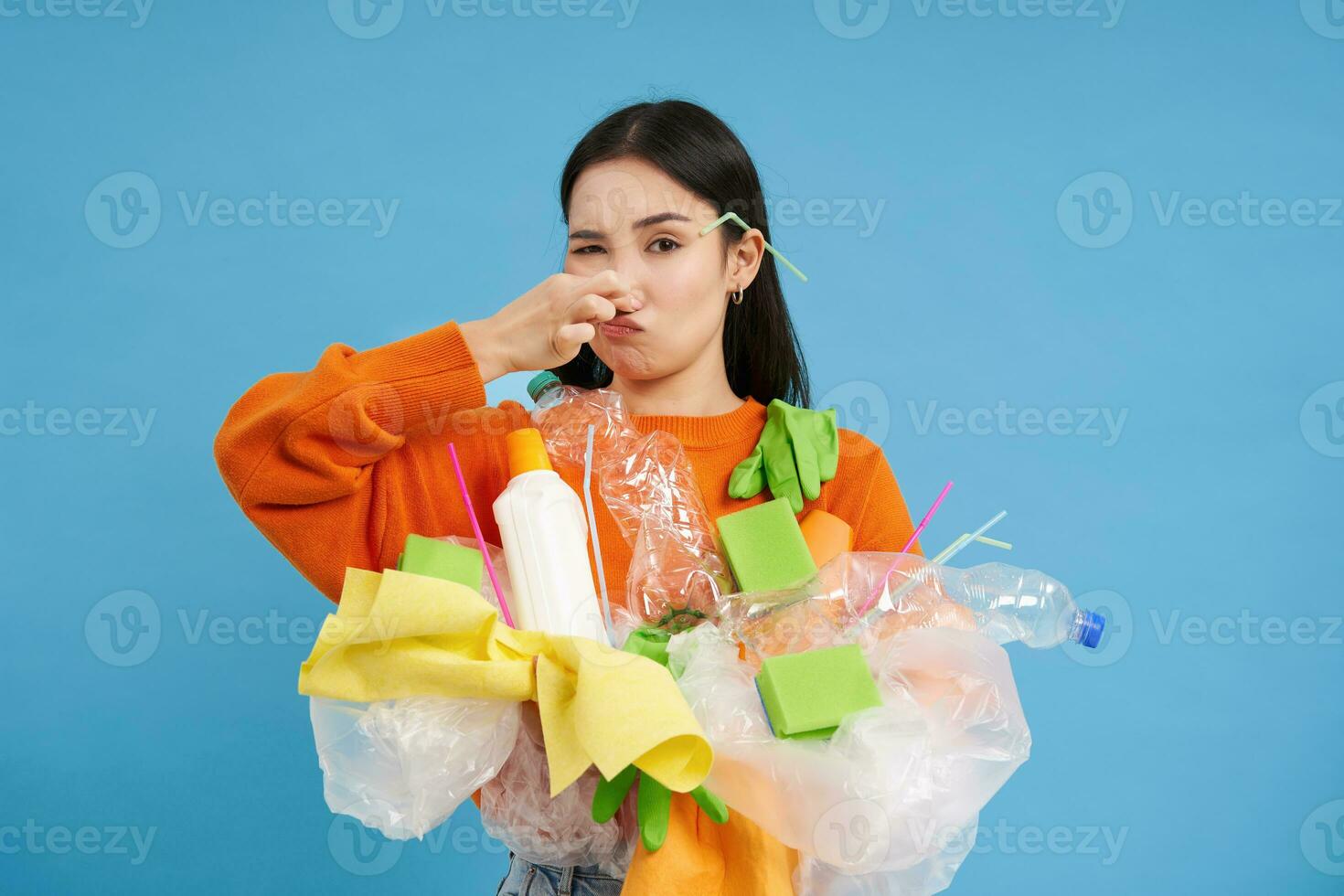 Disgusted asian woman, holds stinky garbage, recycling her waste with disappointed, reluctant face, blue background photo