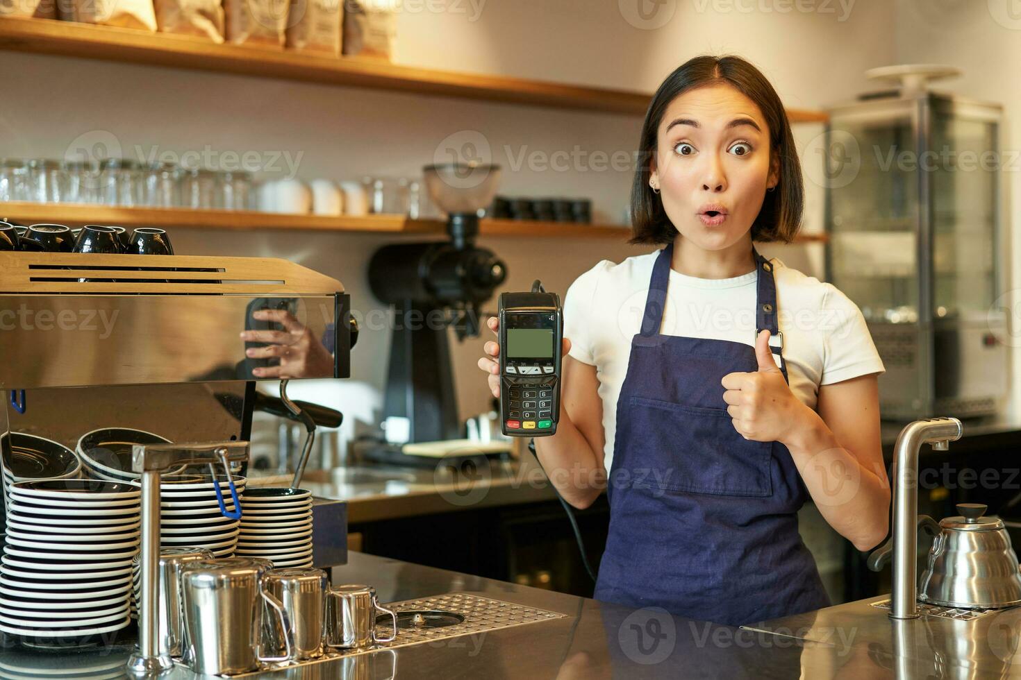 Portrait of cafe barista, girl shows thumbs up and card reader paying machine, recommends contactless payment, looks surprised, stands at counter in apron photo