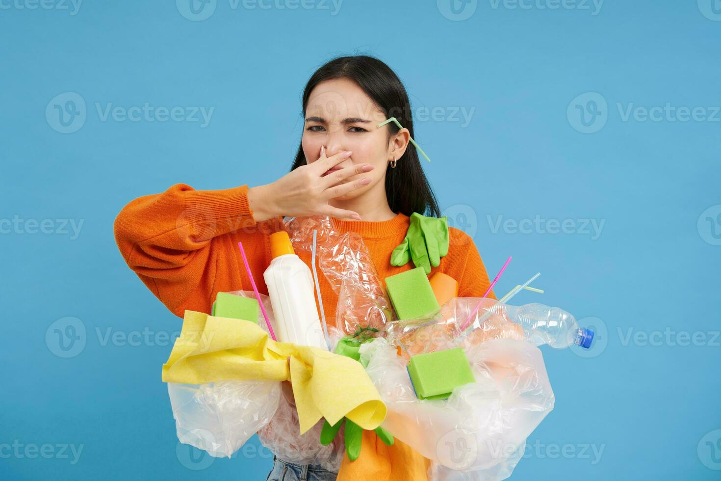Disgusted asian woman, holds stinky garbage, recycling her waste with disappointed, reluctant face, blue background photo