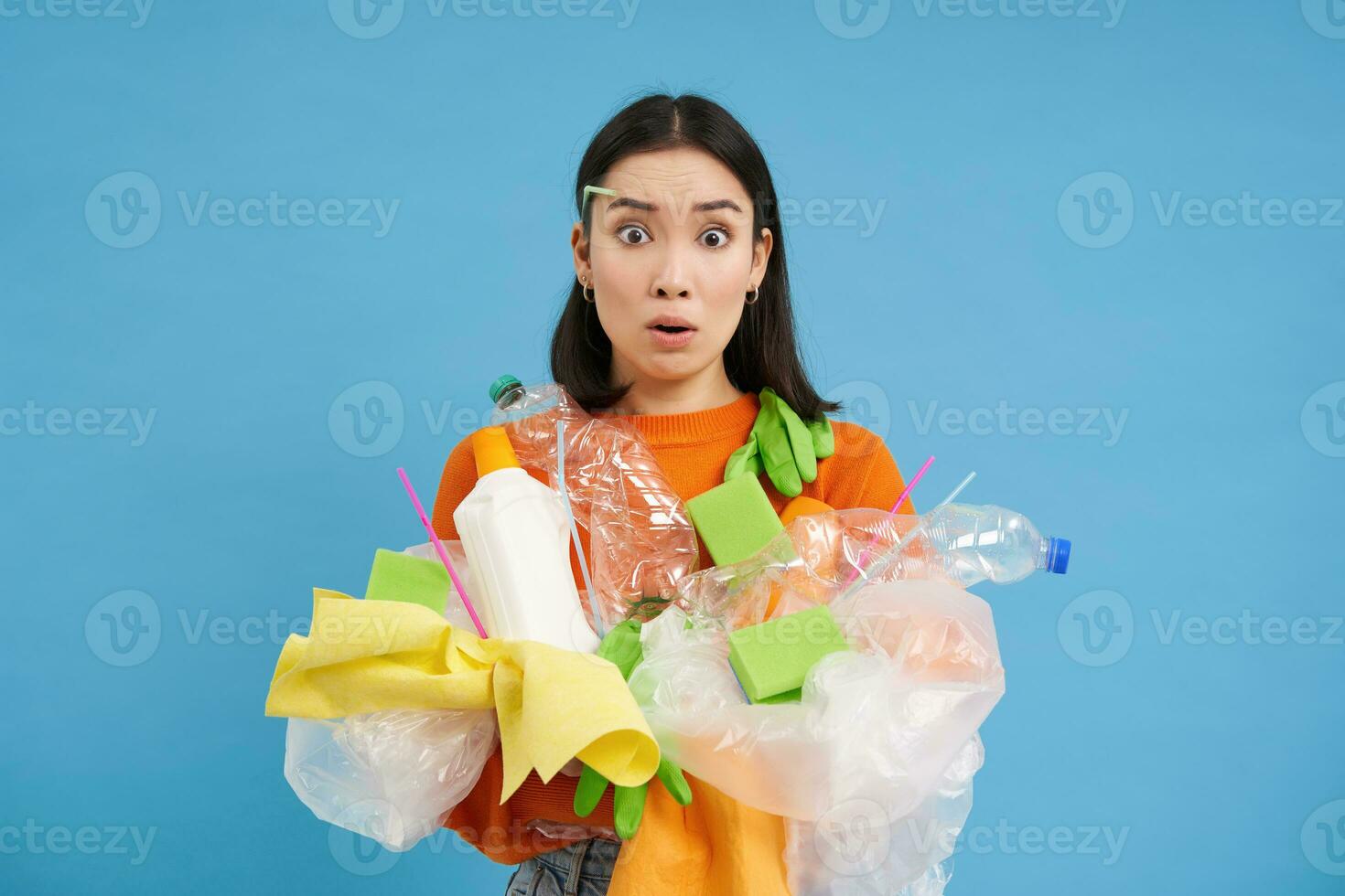 Young confused asian girl with different types of plastic, learns how to recycle, saving enviroment, sorting garbage, blue background photo