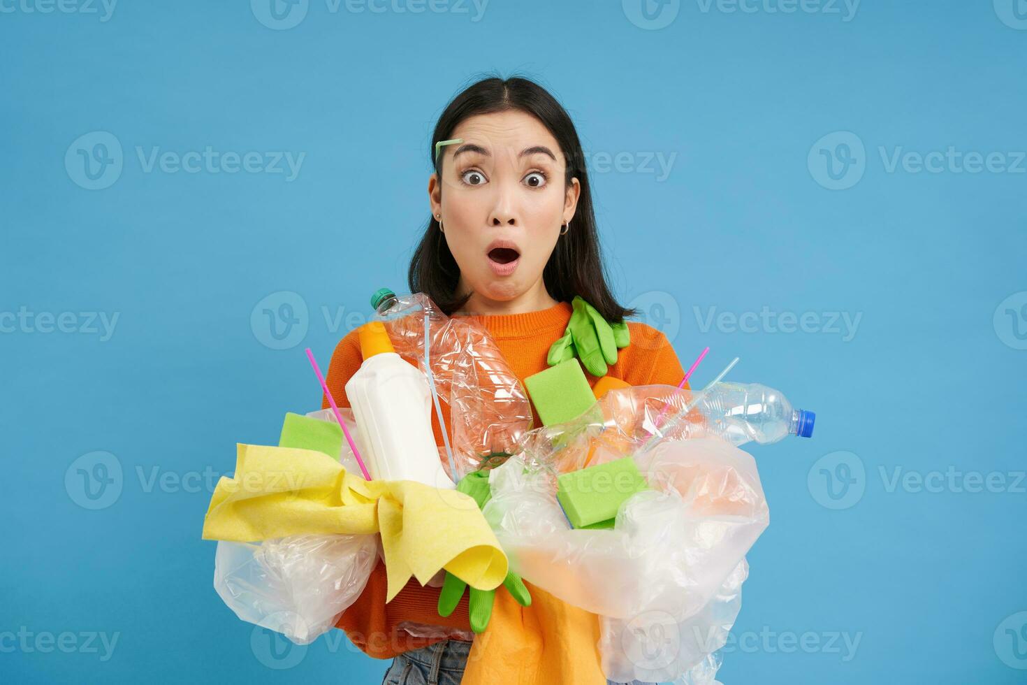Young korean woman looking shocked at plastic waste, holding garbage for recycling with surprised emotion, blue background photo