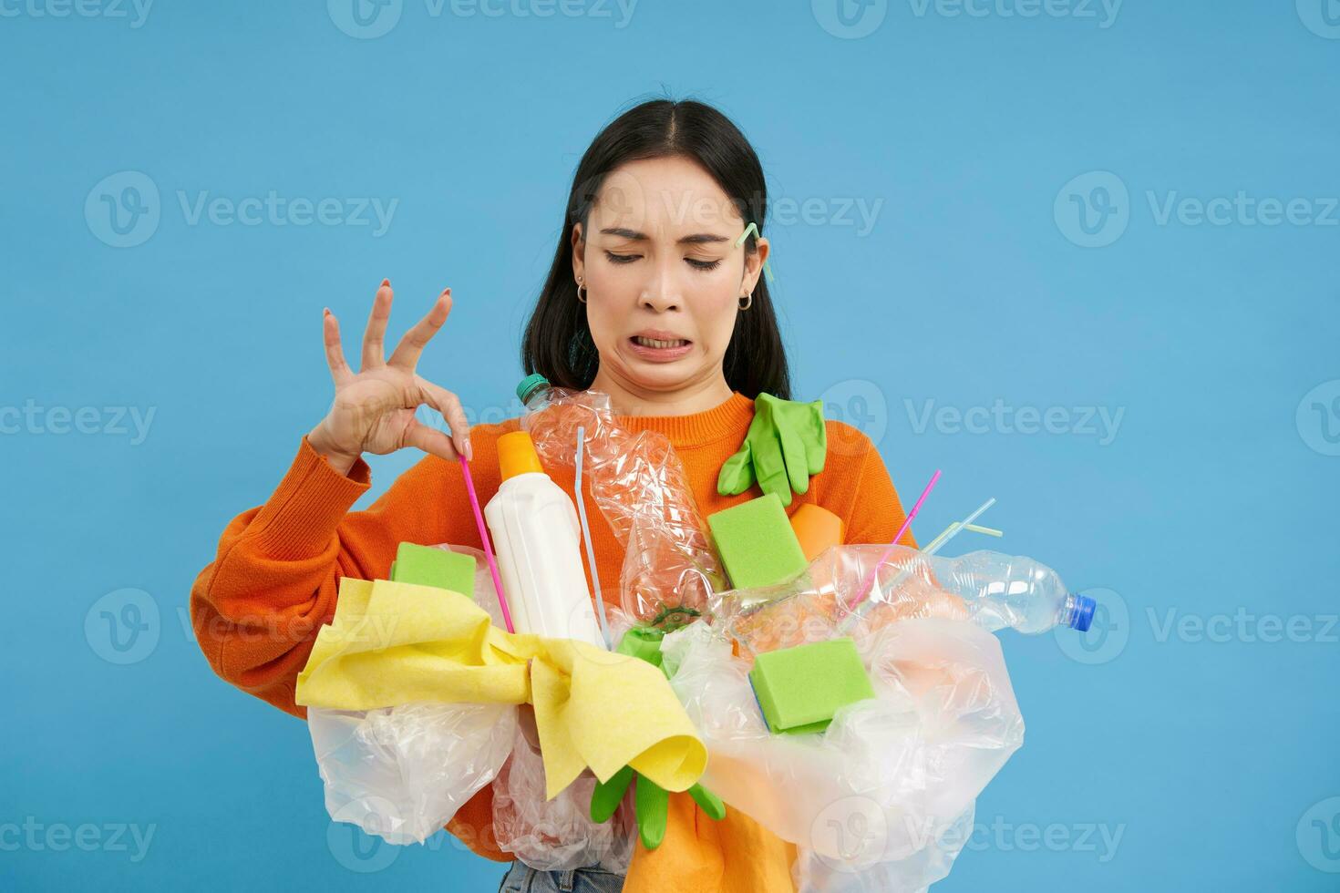 Disgusted asian woman, holds stinky garbage, recycling her waste with disappointed, reluctant face, blue background photo