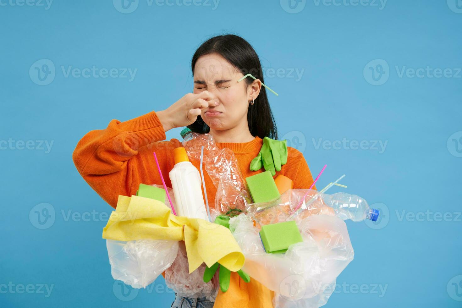 Disgusted asian woman, holds stinky garbage, recycling her waste with disappointed, reluctant face, blue background photo
