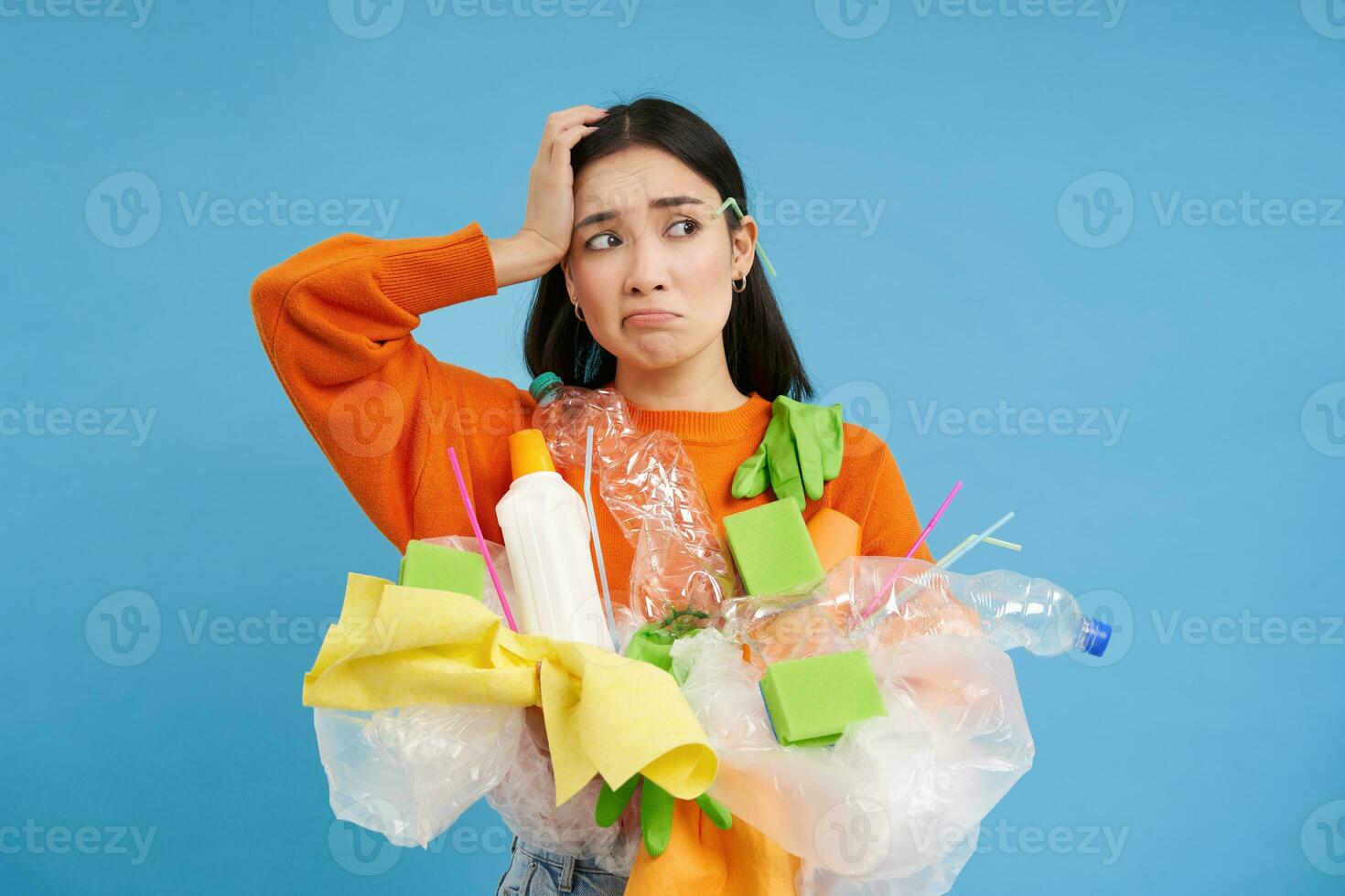 Woman with complicated face, looking confused at plastic garbage, clueless how to recycle items, go green, standing over blue background isolated photo
