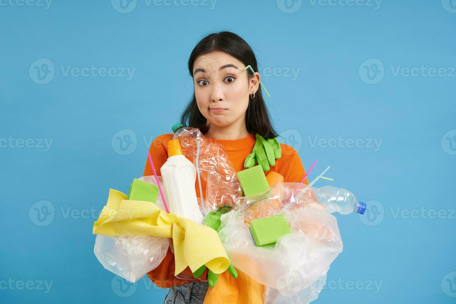 Cute asian woman holding plastic waste, garbage for recycling, looking clueless and shrugging, blue background photo