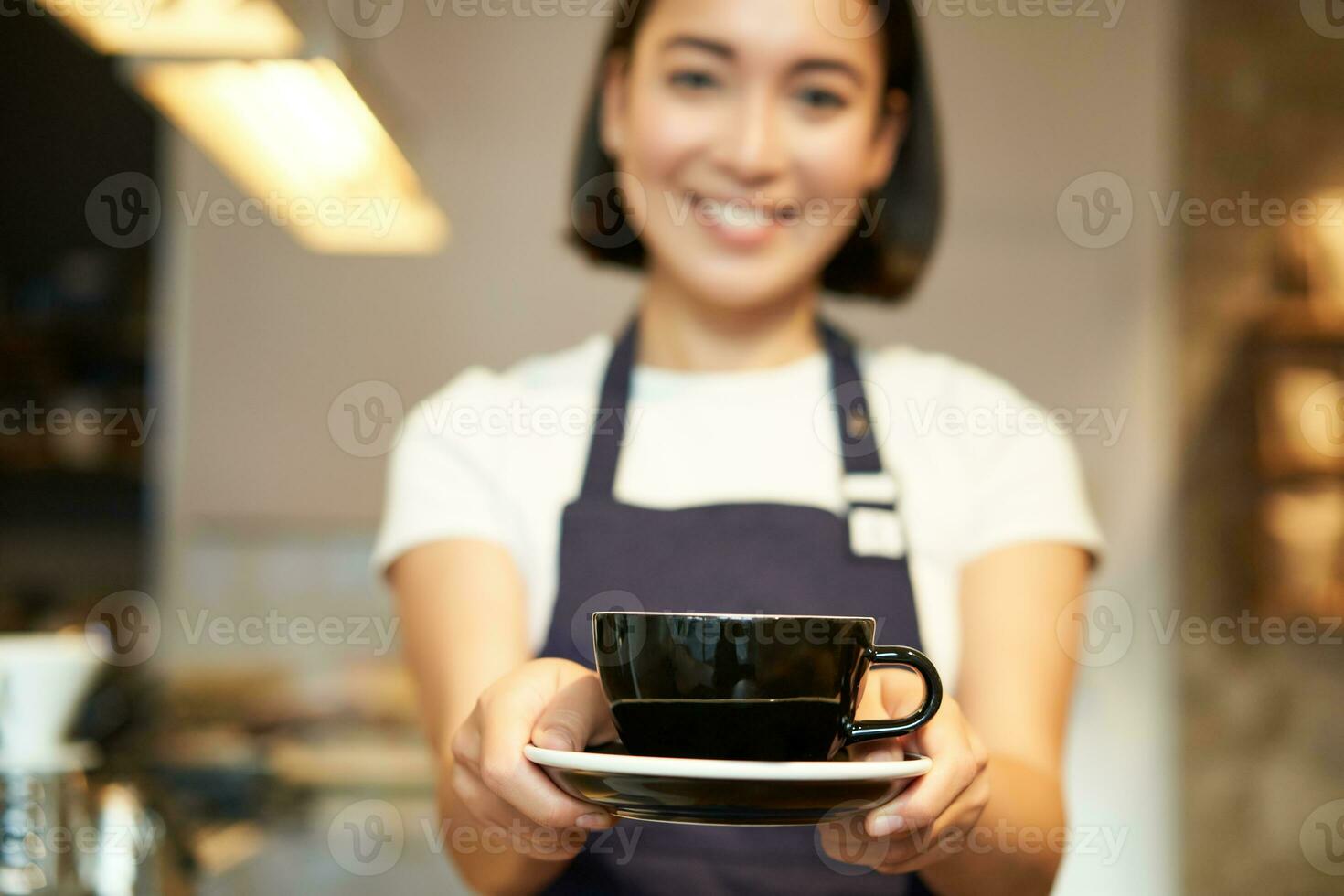 Smiling asian girl barista, standing in apron uniform, giving you cup of coffee, made a drink for client in cafe photo