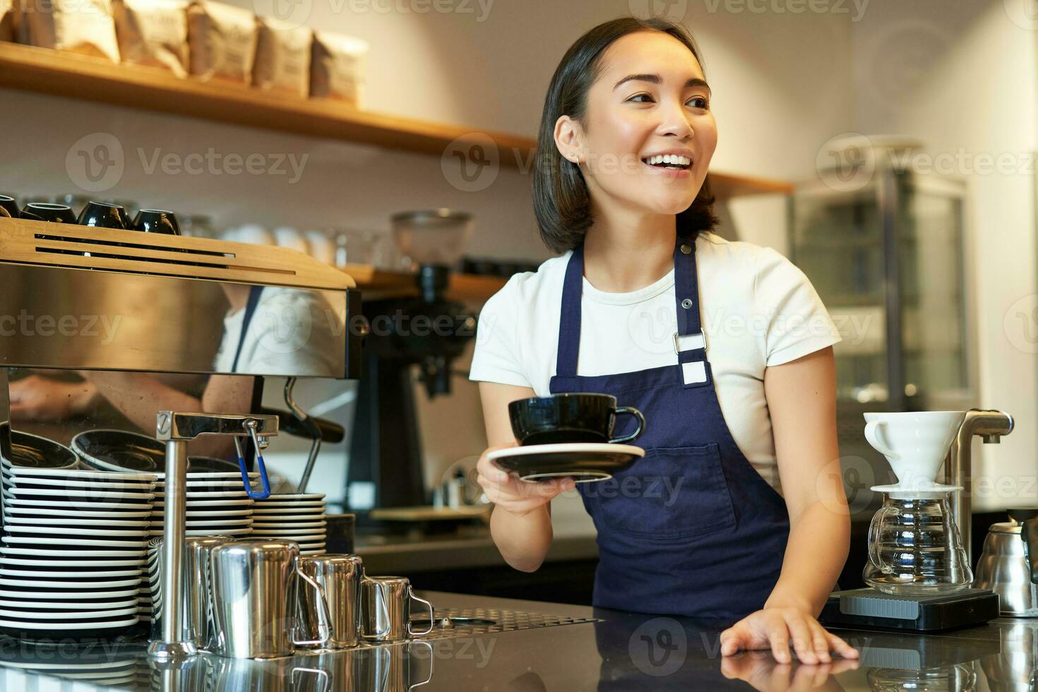 Portrait of beautiful asian girl, student working part-time in cafe, holding cup of coffee, made order, looking for client, wearing uniform photo