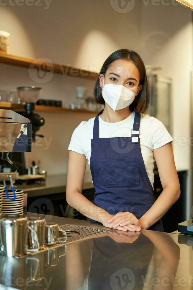Vertical shot of friendly barista, female staff member in medical mask, working behind counter with clients, serving coffee in cafe, standing in uniform apron photo