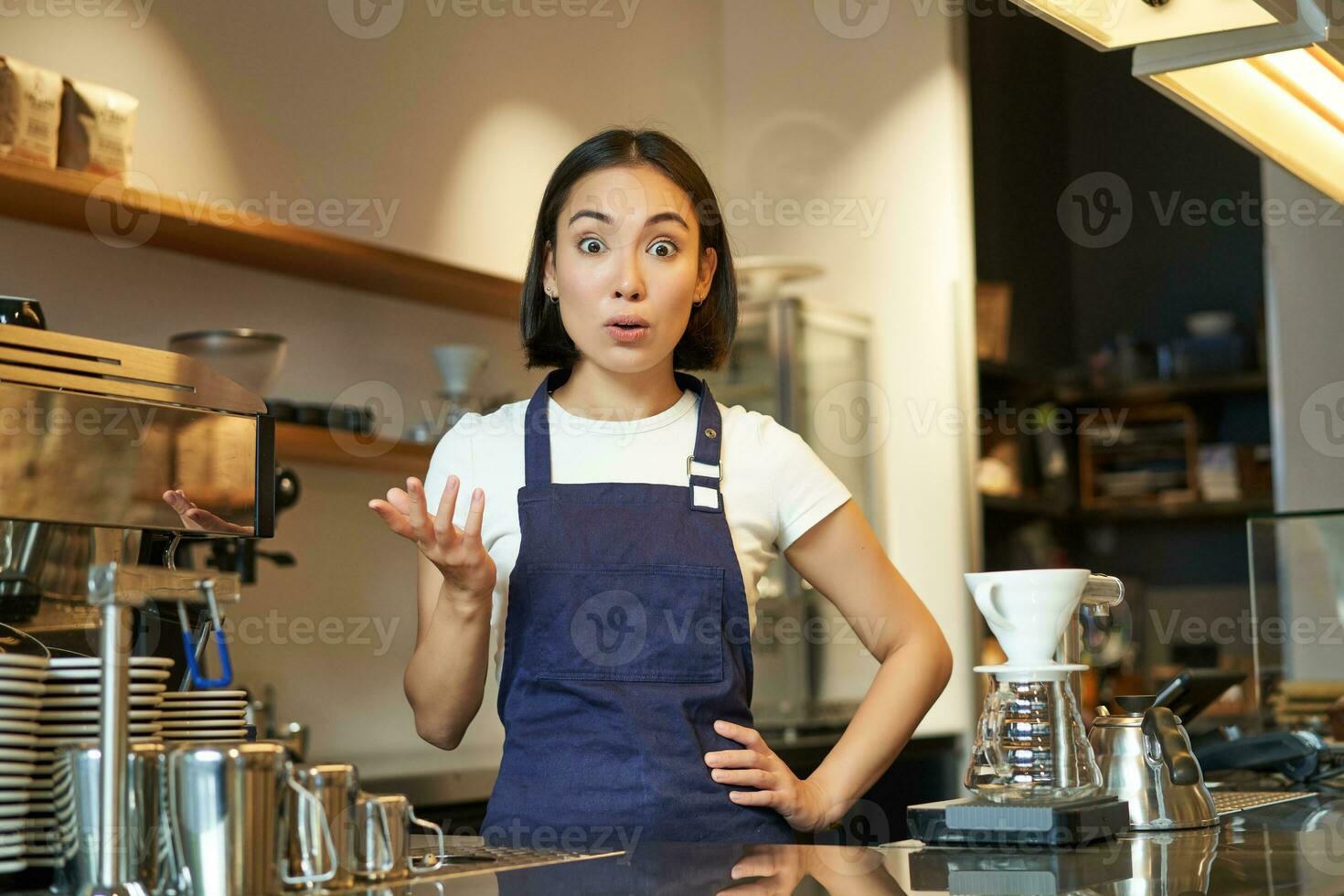 retrato de niña barista en delantal, mira sorprendido, puntos con mano y miradas asombrado a cámara, trabajos detrás mostrador y brebajes café foto