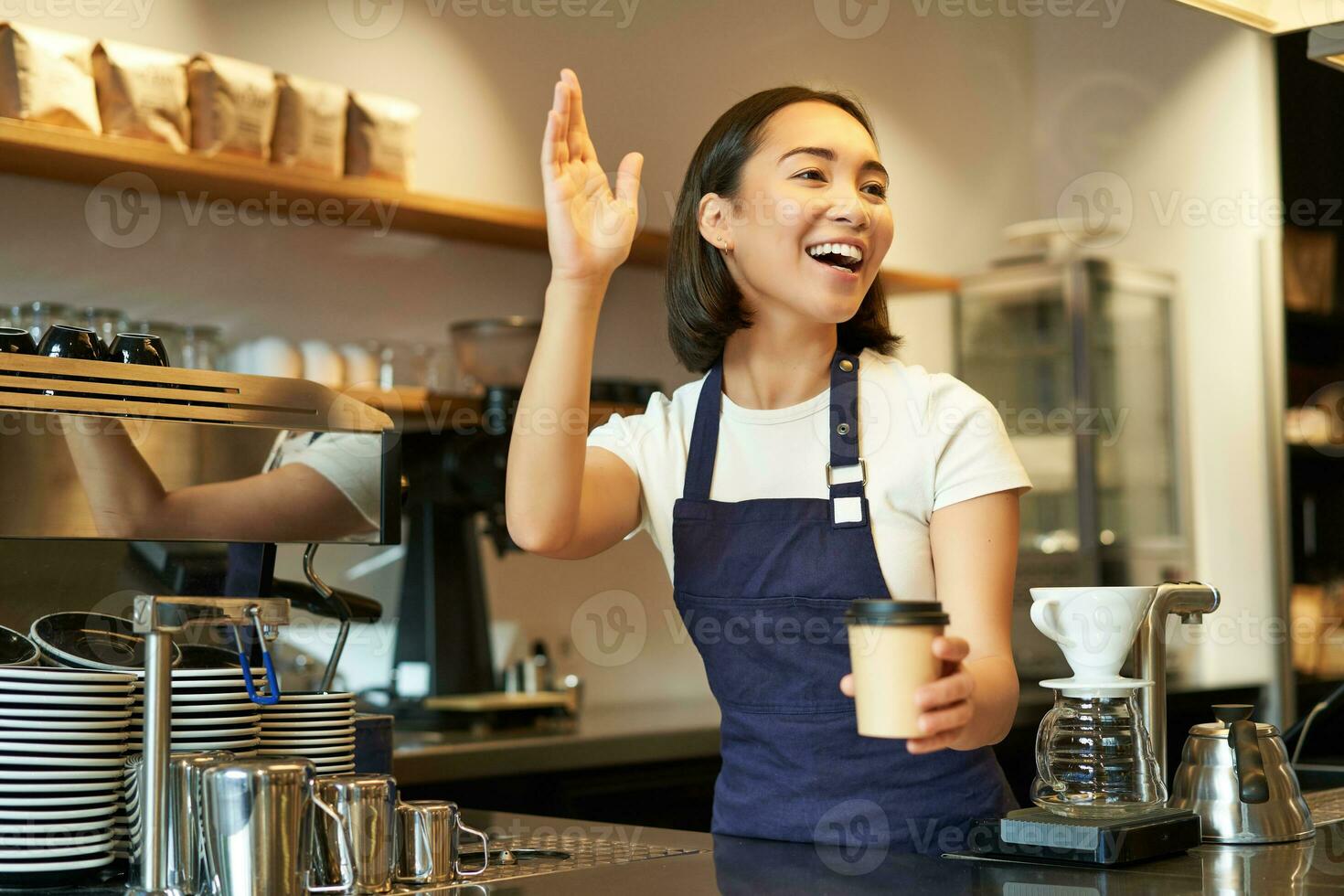 Friendly smiling barita girl waving at client, holding their cup of coffee, prepared takeaway order, working in cafe behind counter photo