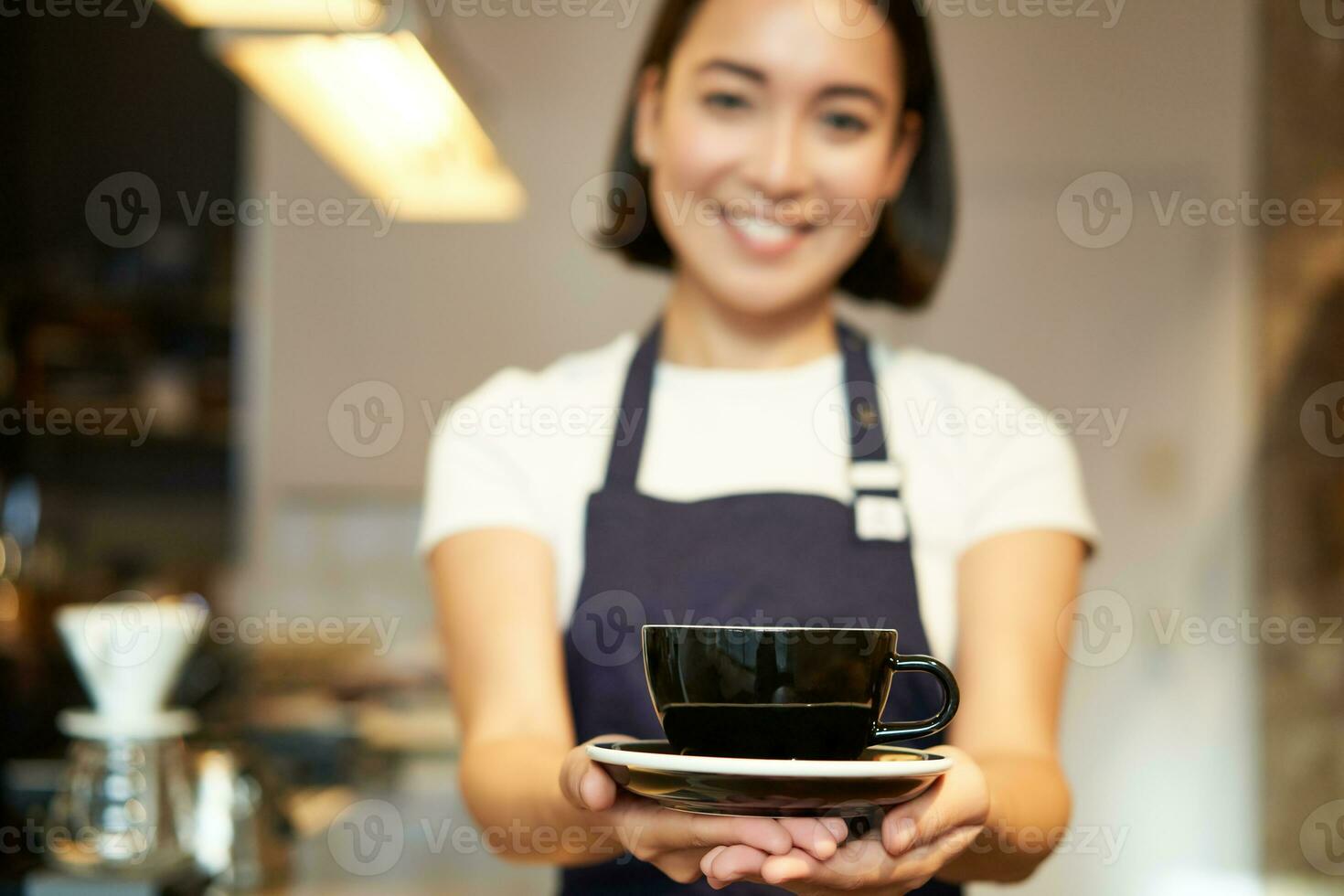 retrato de sonriente asiático niña barista, camarera en café uniforme, dando usted taza de café, preparar bebida para cliente, mirando simpático foto