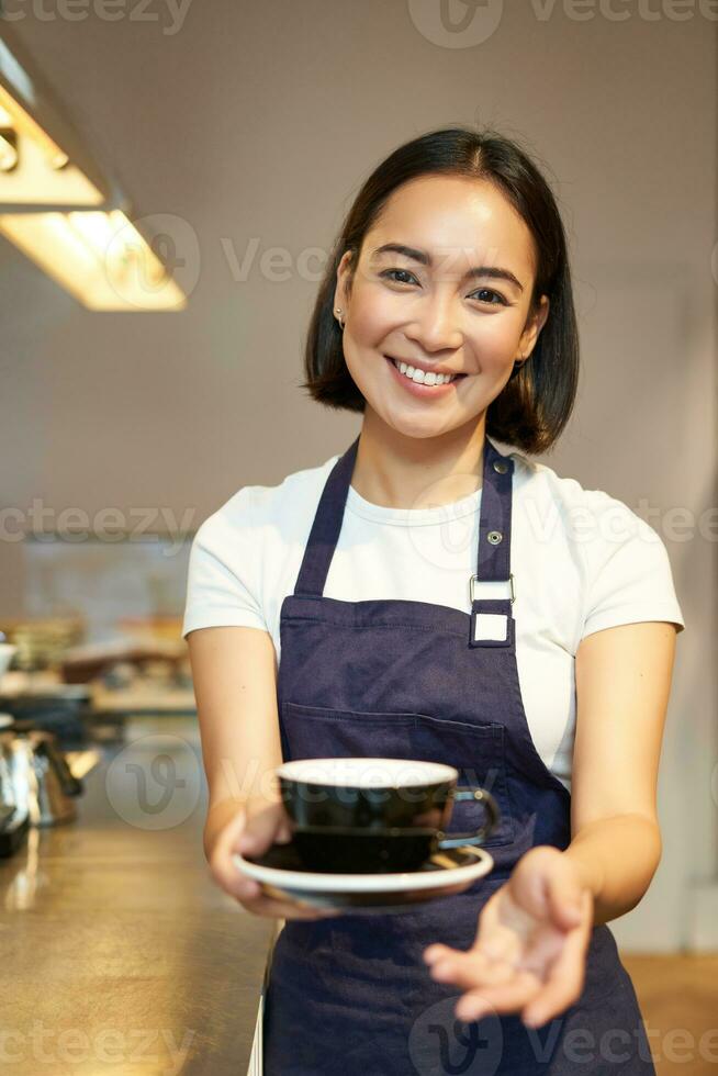 Smiling korean girl in uniform, working in cafe, giving you cup of coffee, preparing order for client photo