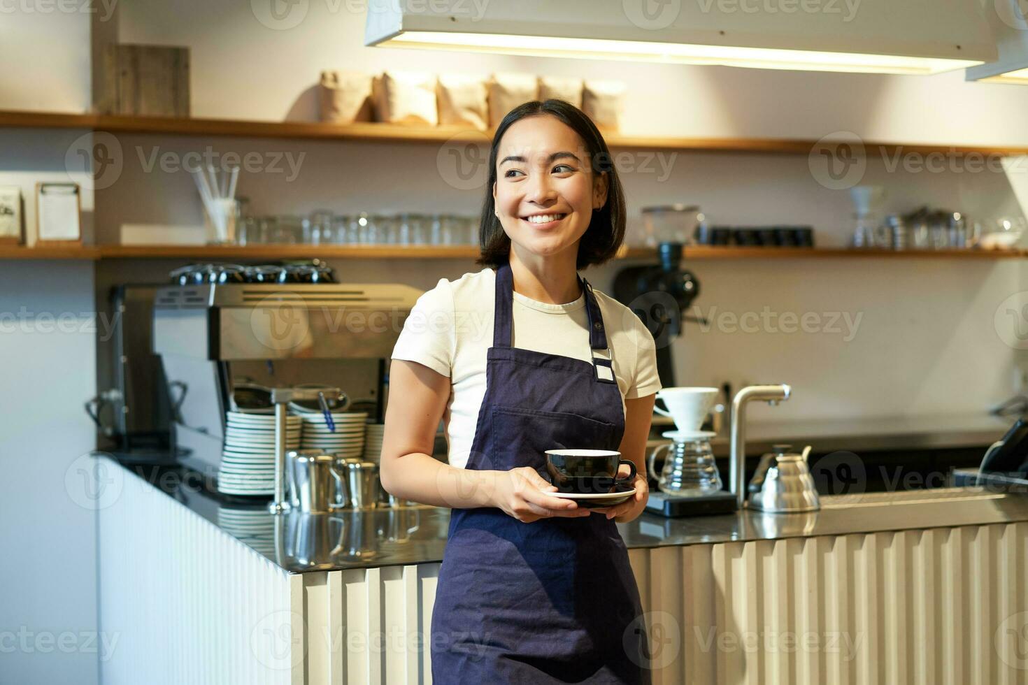 Portrait of asian smiling female barista, cafe waitress holding cup of coffee, serving clients, taking order to the tablet, standing near cafe counter photo