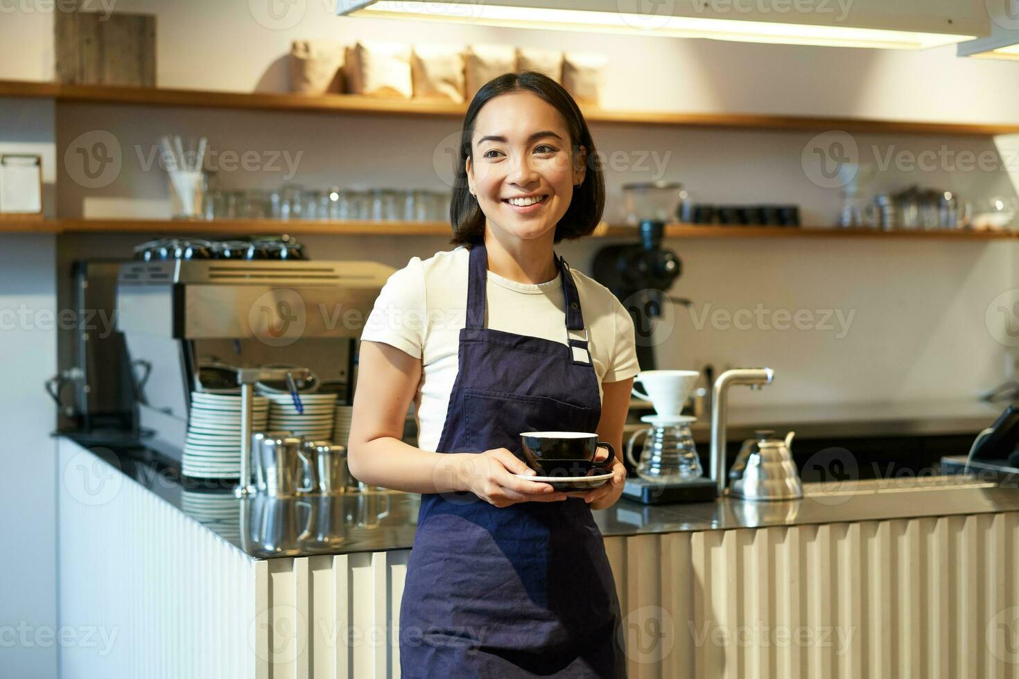 retrato de asiático sonriente hembra barista, café camarera participación taza de café, servicio clientela, tomando orden a el tableta, en pie cerca café mostrador foto