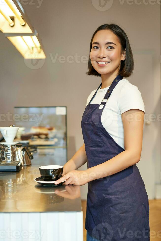 Vertical shot of smiling asian girl barista, wearing uniform, making coffee, standing near counter with cup, working in cafe photo