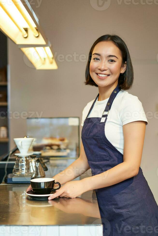 Vertical shot of smiling asian girl barista, wearing uniform, making coffee, standing near counter with cup, working in cafe photo