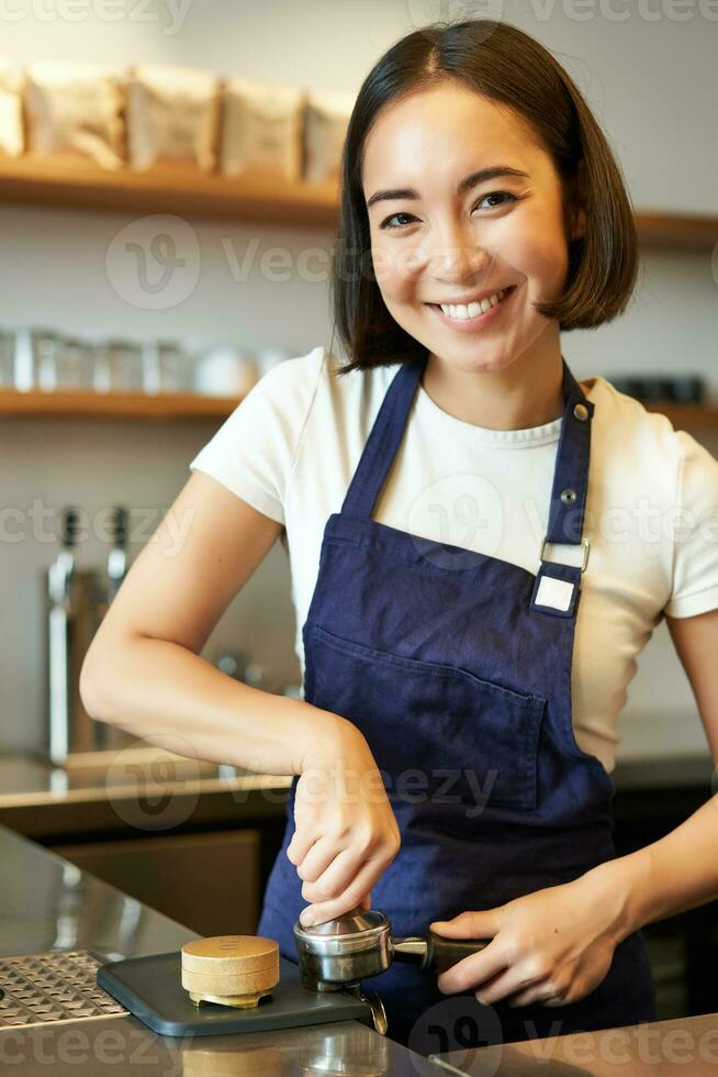 Vertical shot of barista, asian girl in cafe using tamper for coffee machine, wearing blue apron, preparing order photo